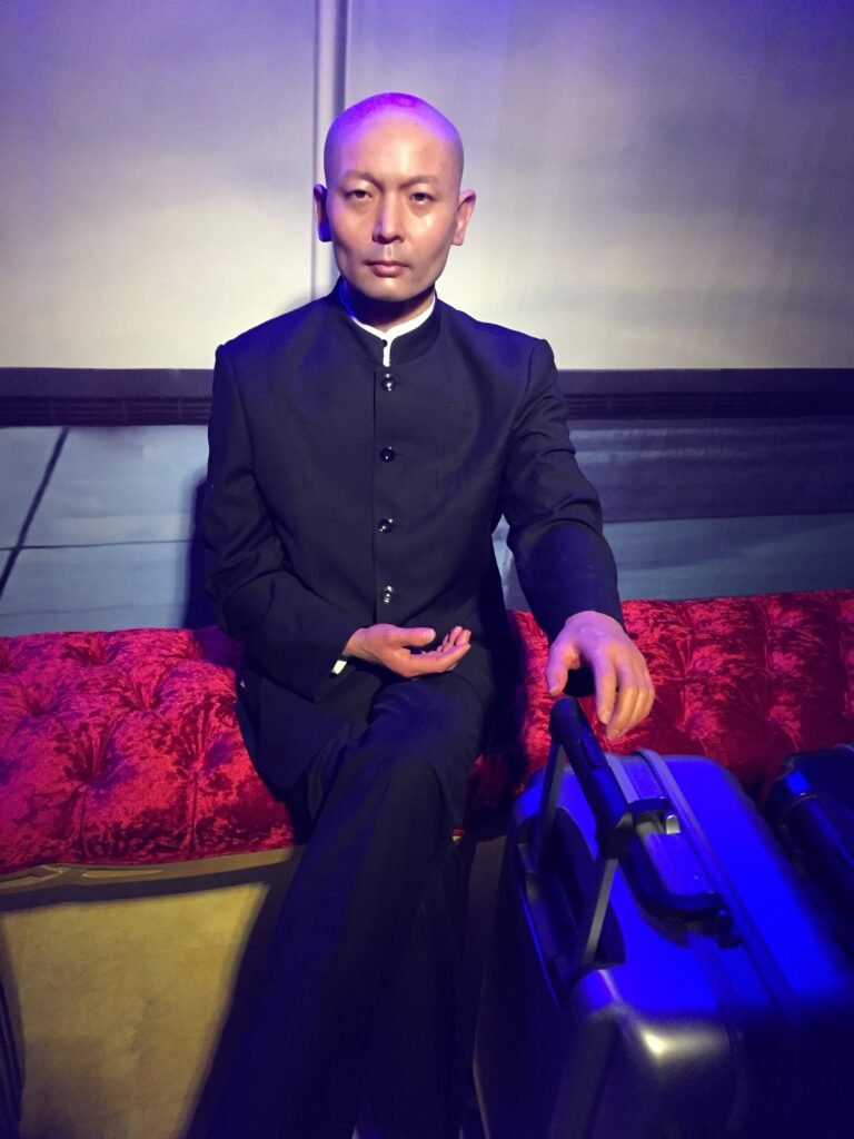 Ge You wax figure seated on a red velvet sofa in a museum setting.