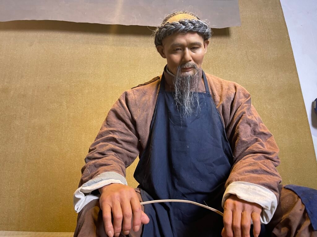 Full seated view of traditional craftsman wax figure weaving a bamboo basket in a cultural exhibition setting.