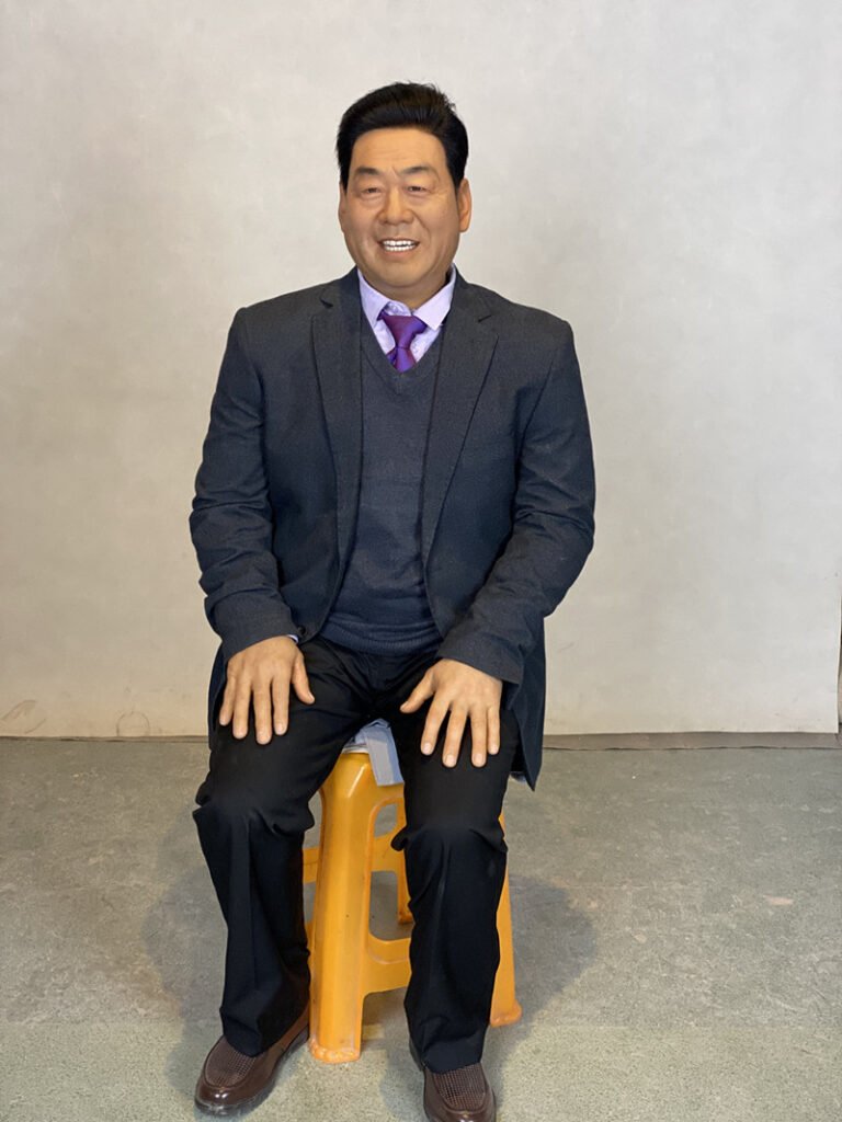 Full seated view of contemporary business leader wax figure in tailored suit for exhibition display.